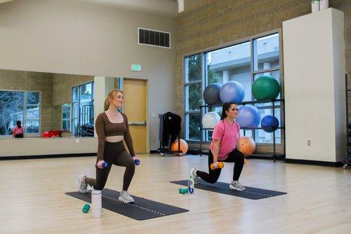 Two women performing lunges on yoga mats in a fitness studio.